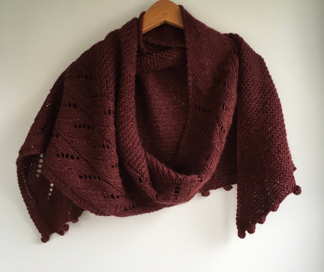 maroon colored shawl with cables and bobbles draped on wooden hanger