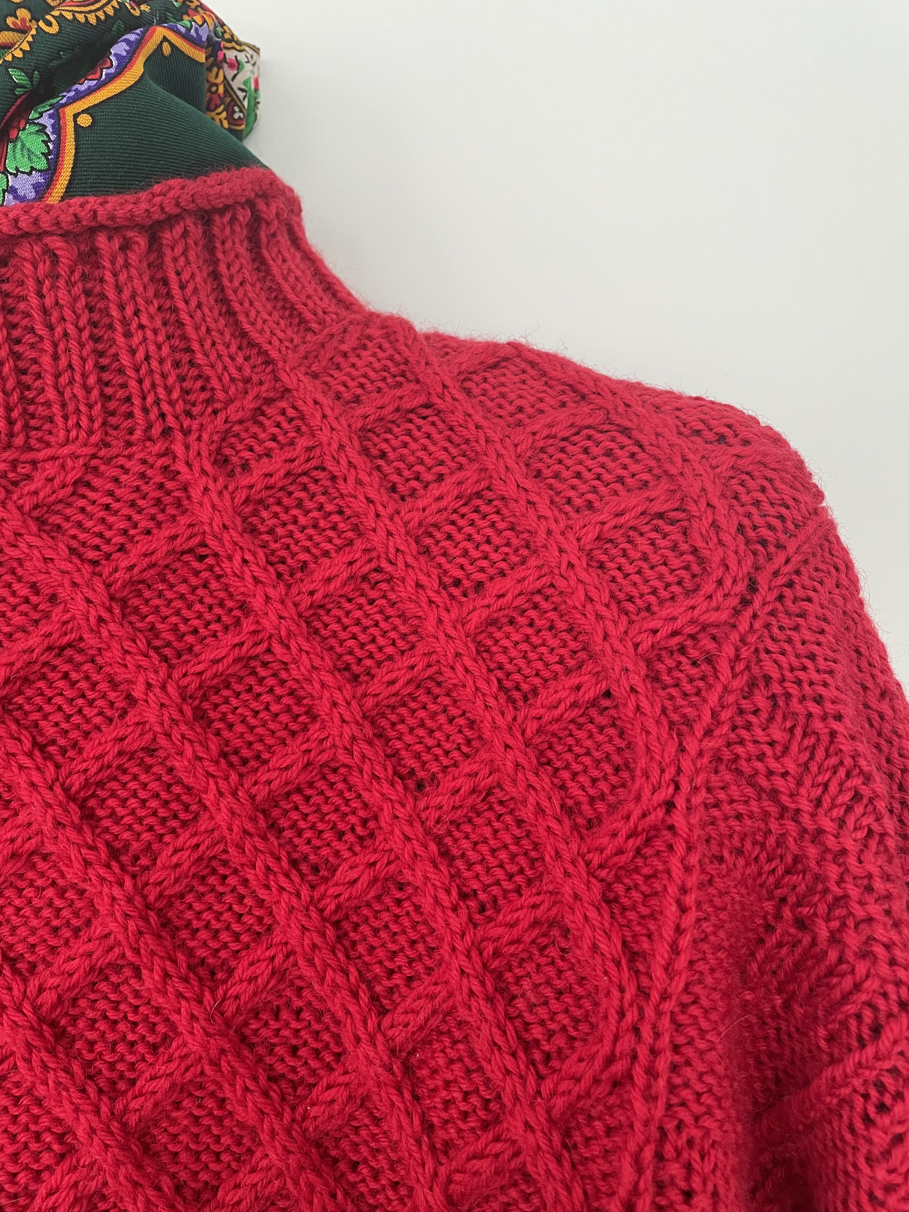close in detail of red hand knit sweater showing cable front and ribbed rolled collar and green scarf at the neck