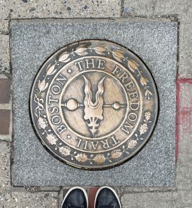 bronze medallion marking Boston Freedom Trail set in sidewalk with the tips of two black leather shoes on bottom edge