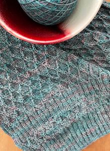 handknit teal shawl with cables and red porcelain bowl holding yarn in upper left corner