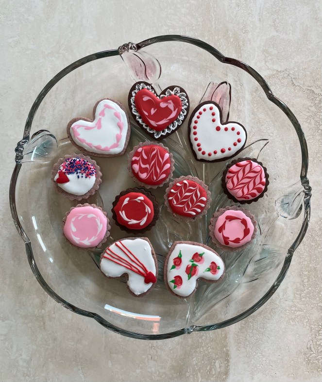 12 round and heart shaped cookies decorated with pink, red and white frosting on a clear glass plate