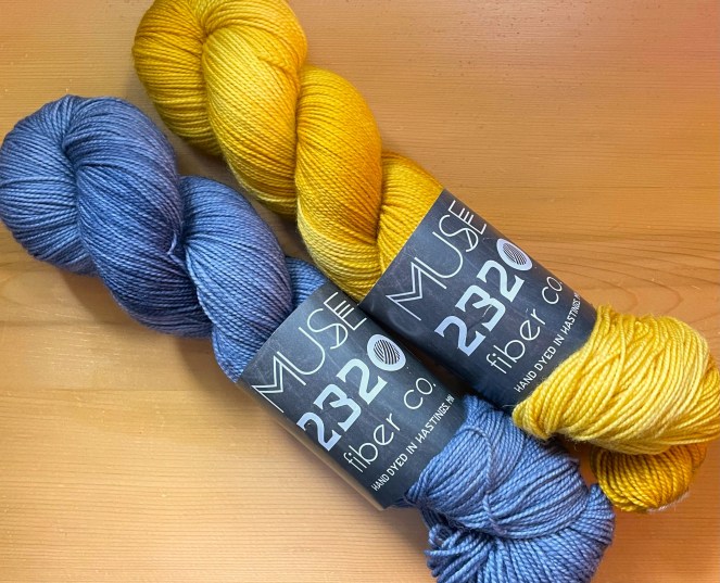 one skein of blue yarn and one skein of gold yarn each with a black Muse2320 paper label
