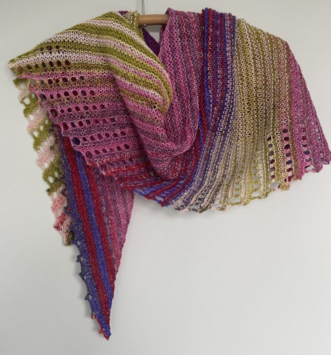 A hand knit lace shawl in multi colored pinks and greens on a wooden hanger
