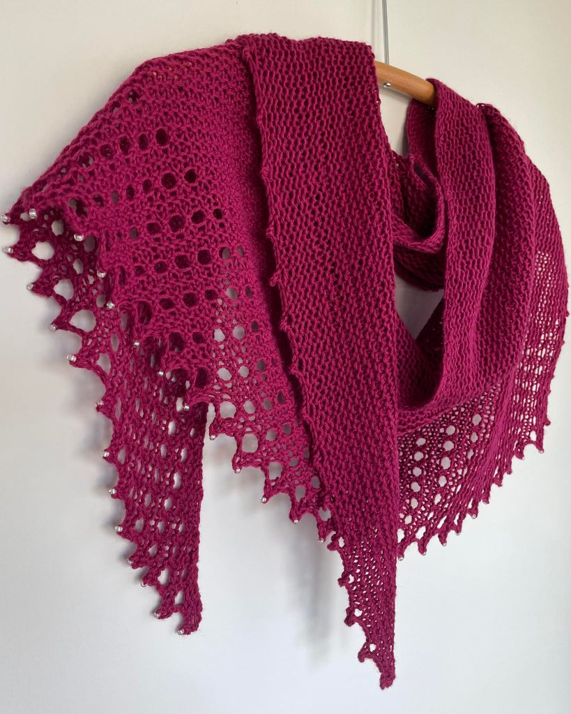 hand knit fuchsia colored shawl with lace edges and crystal beads on a wooden hanger