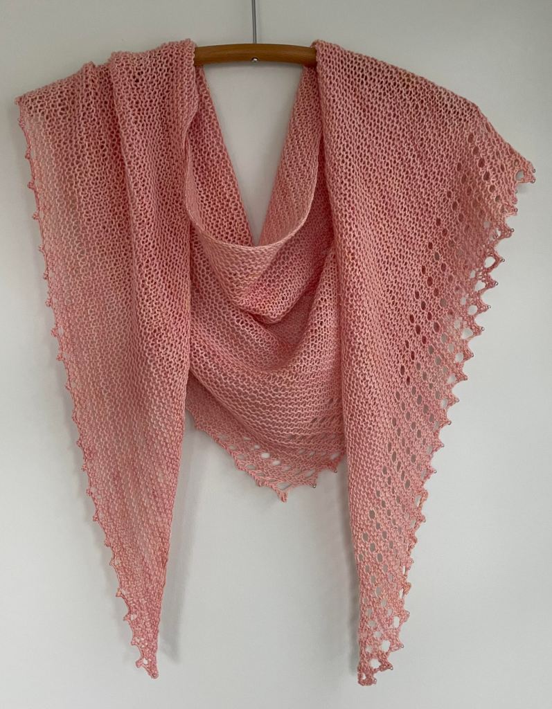 hand knit peach colored shawl with lace edges on a wooden hanger