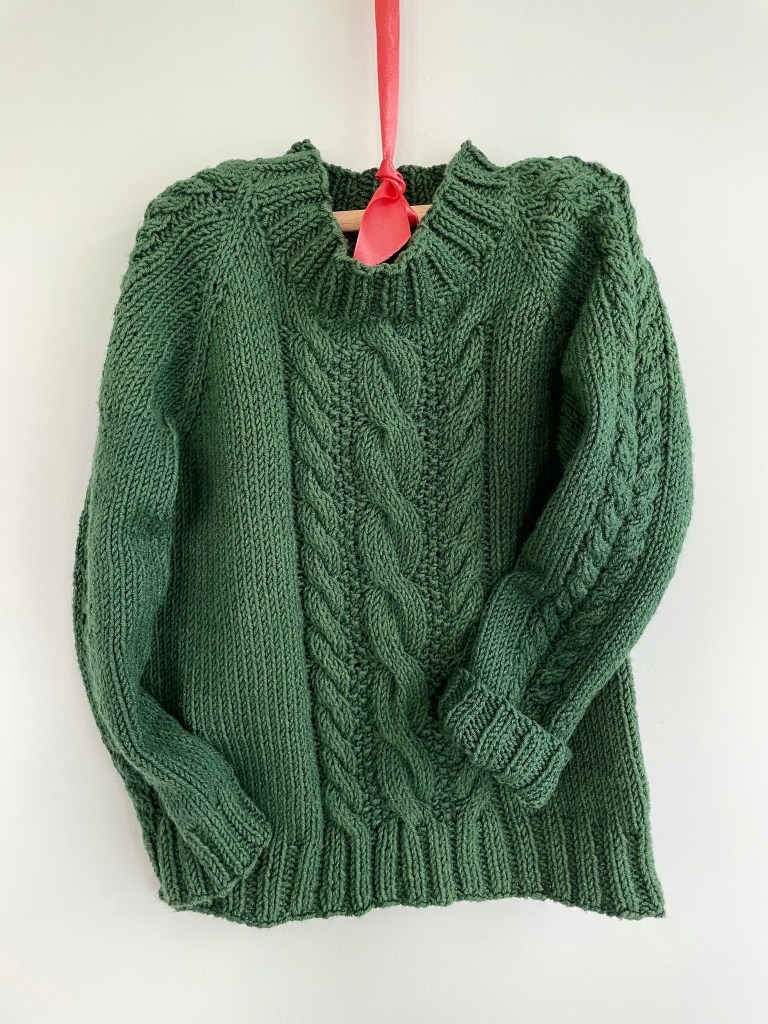 handknit green sweater on white background with a central cable design on the body and the sleeve hanging on a wooden hanger suspended by an orange ribbon