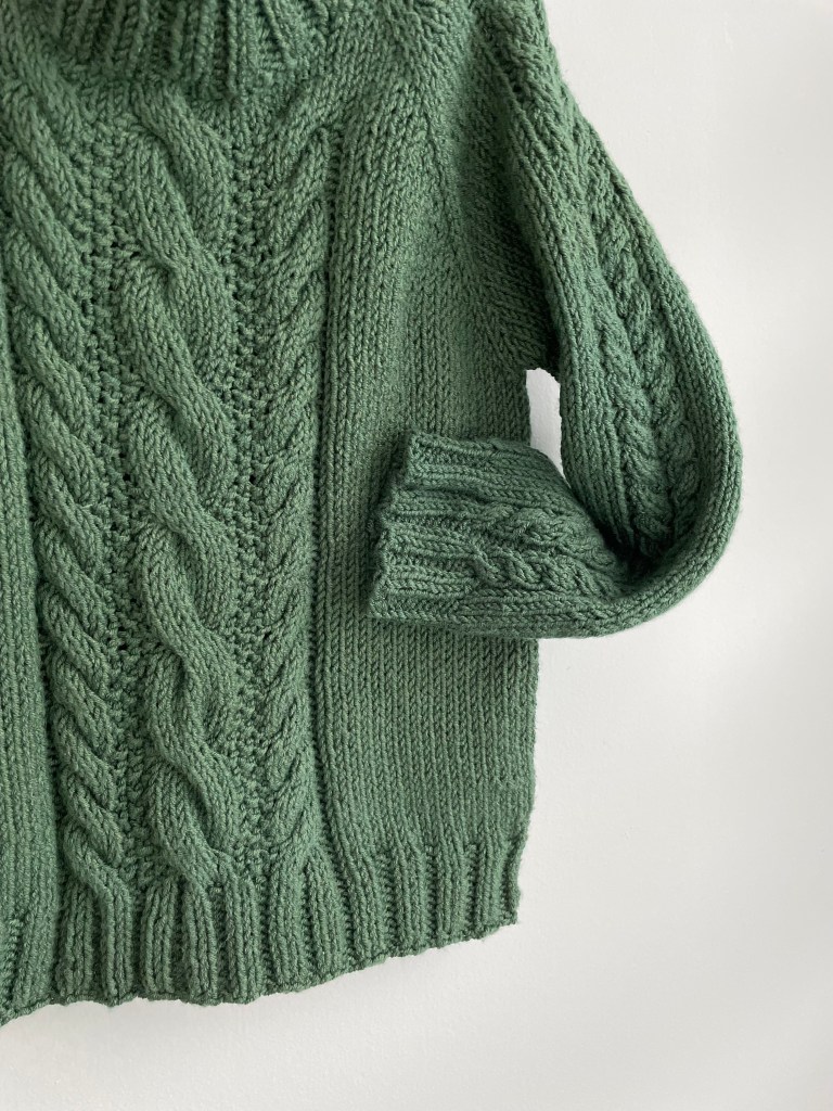 details of a handknit green sweater on white background with a central cable design on the body and the sleeve