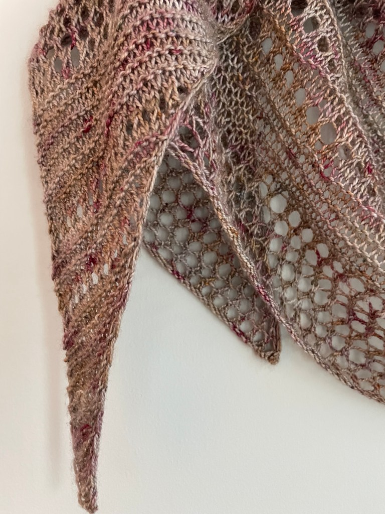 close up detail of handknit shawl with rows of lace stitches in lavendar and green hues on a wooden hanger