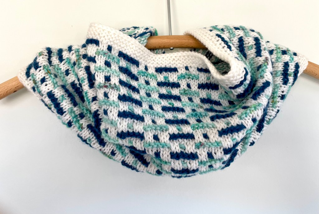 hand knit cowl with white background and blue and teal dashes of color displayed on a wooden hanger