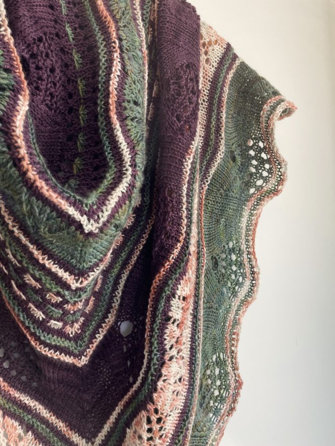 close up detail of a handknit shawl in purple, green, and peach colors on a white background