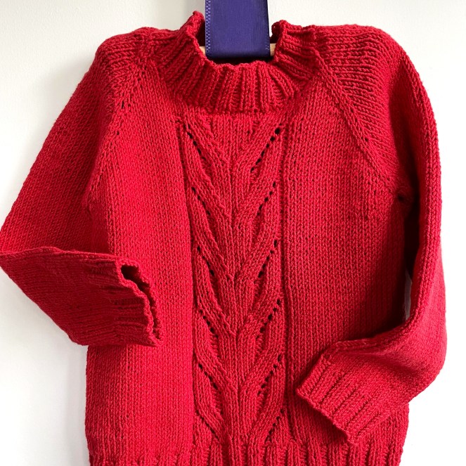 handknit red sweater with front cable design hanging from a purple ribbon