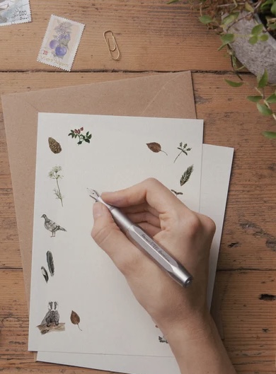 hand holding pen poised above stationary on a wooden table; image from the Sunday Letter Writing Project